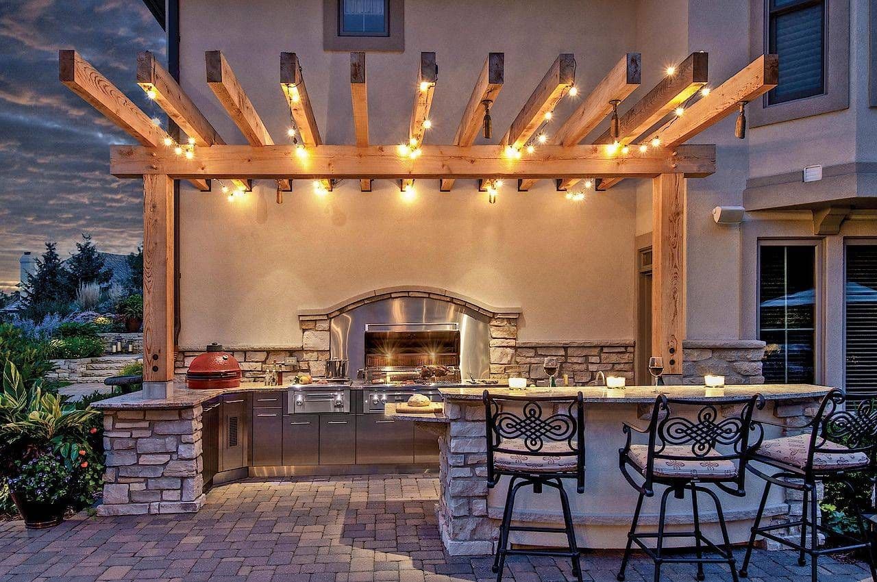 Outdoor stone-and-wood patio kitchen glowing with string lights, built-in grill, bar seating, and ornate stools at dusk.