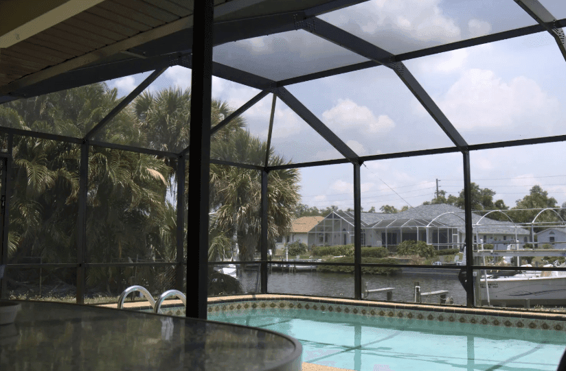 Screened backyard pool overlooks a canal and neighboring homes, framed by palm trees beneath a sunny, partly cloudy sky; calm tropical setting.
