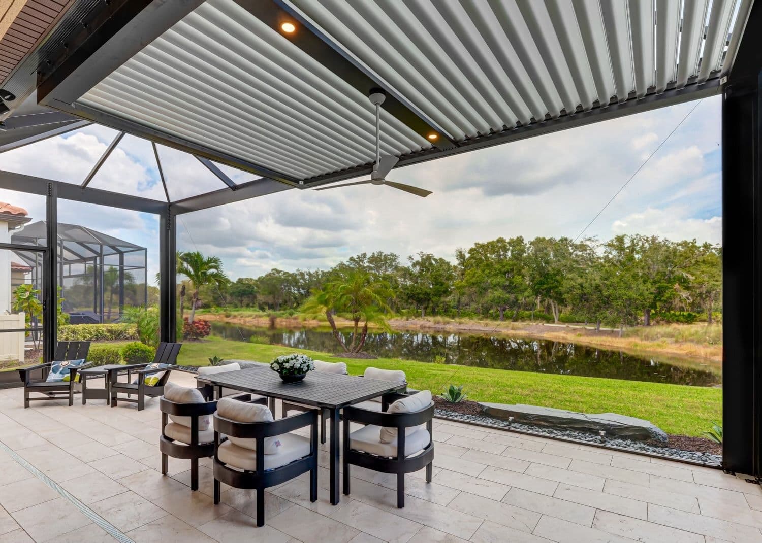 Covered patio with modern seating, a fire pit, and potted plants. Overhead slatted pergola provides shade. Greenery and open sky create a serene atmosphere.