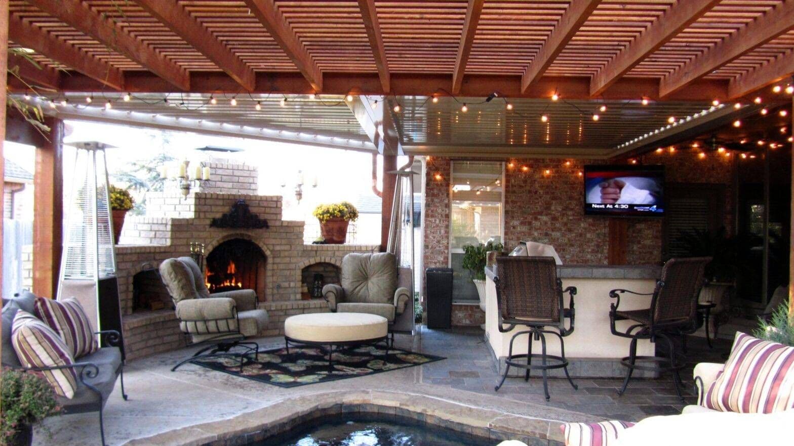 Covered outdoor patio with wood pergola, string lights, stone fireplace, cushioned seating, bar table, TV, and a warm, inviting atmosphere.