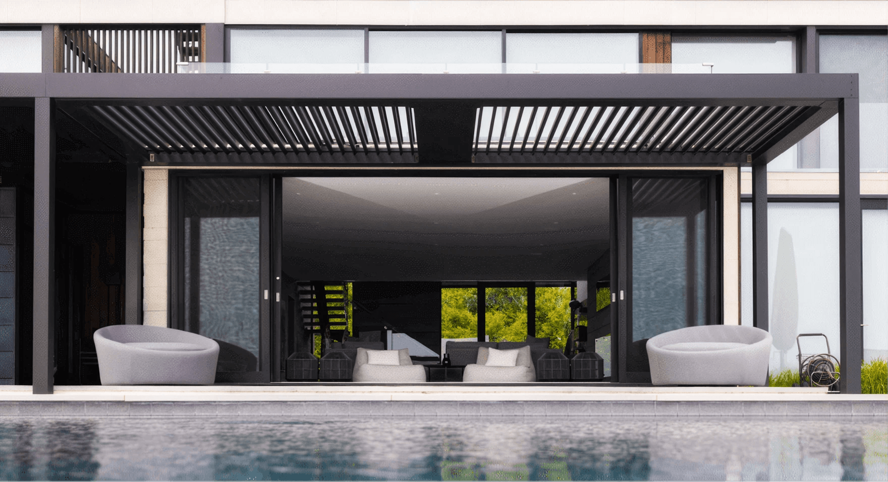 Modern luxury home with a covered patio with a pergola, open glass doors, white lounge chairs, indoor seating, lush greenery, and a calm pool.