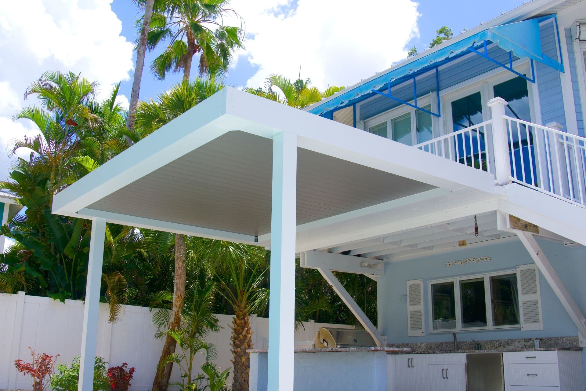 Bright tropical home patio with a white, closed, louvered pergola, palm trees, outdoor kitchen, and balcony beneath a sunny, cloud-filled sky.