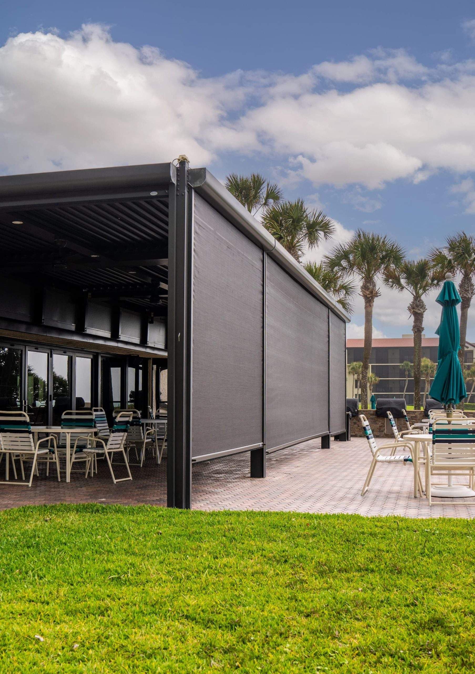 Modern patio with louvered pergola, retractable shade screens, dining furniture under covered structure, and teal umbrella with palm trees in background.