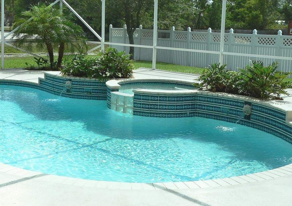 Sunny backyard swimming pool with bright turquoise water, curved patterned tile, tropical plants, and a white fence in a calm, inviting setting.
