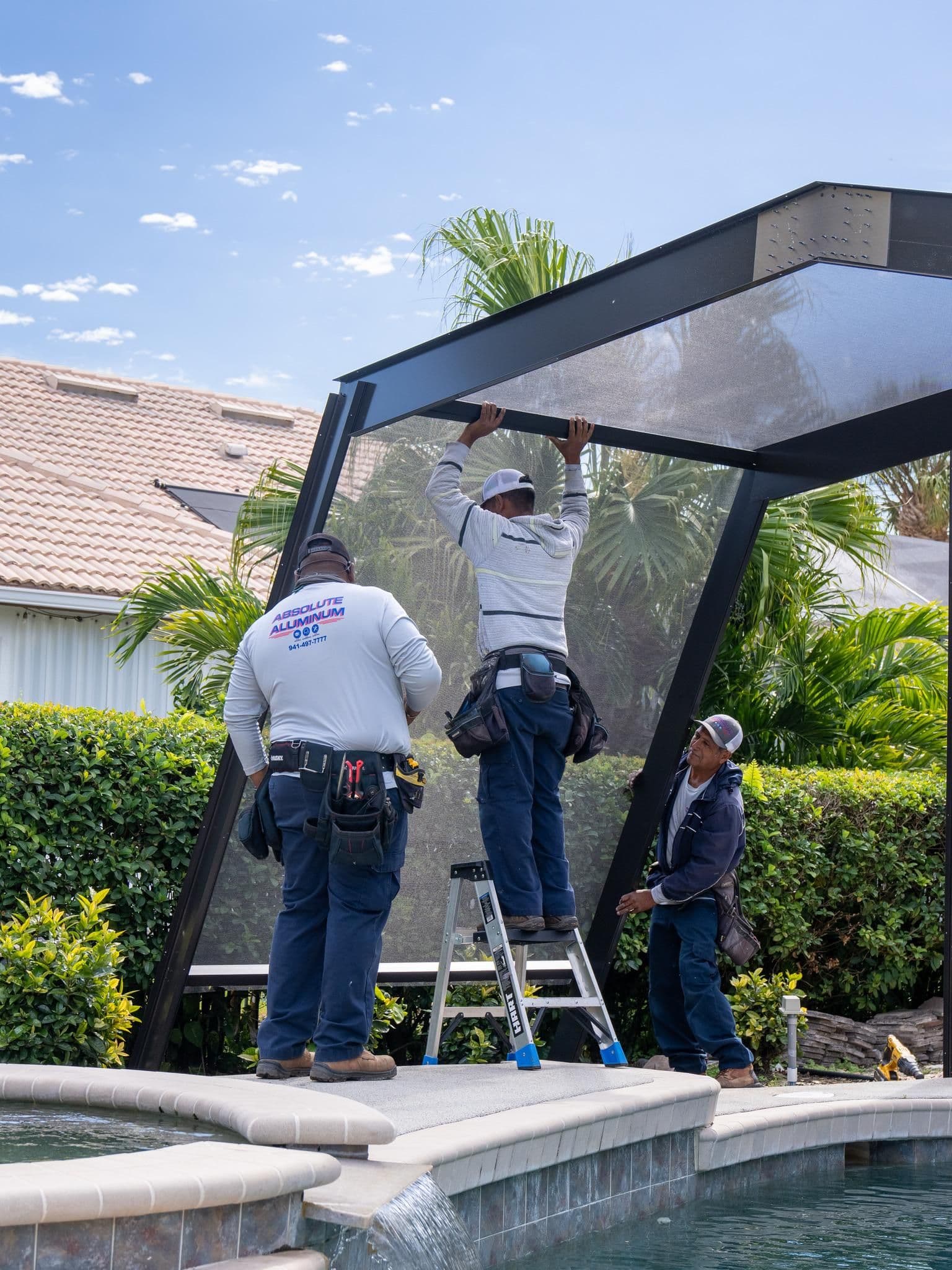 Three workers installing a black aluminum pool cage over a swimming pool in a residential backyard