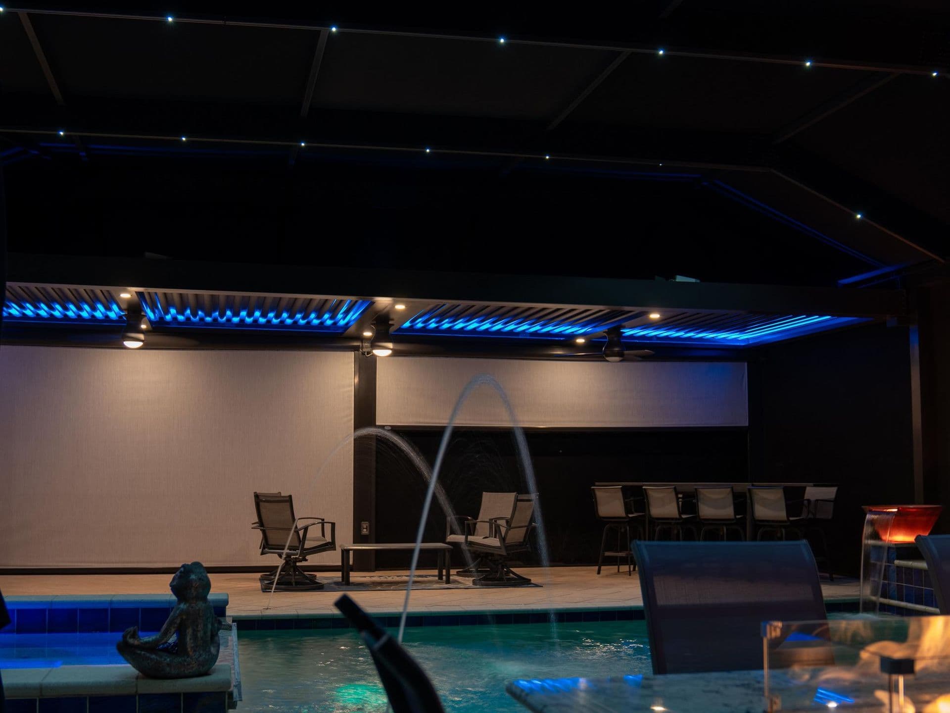Poolside patio at night with blue LED screen enclosure lights, lounge chairs, a projection screen, and pool fountain with underwater lighting