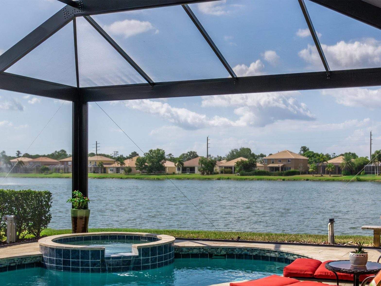 Screened pool patio with turquoise pool, raised spa, red cushions, and a serene lake view of suburban homes beneath a bright blue sky.