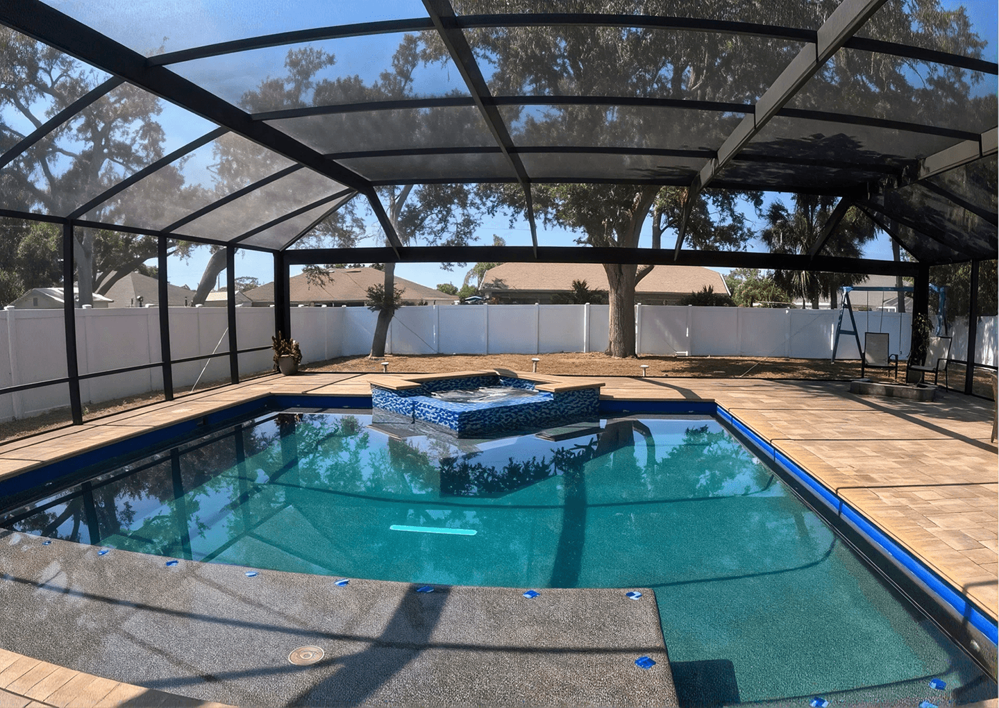 Residential swimming pool with attached hot tub under black screen enclosure, surrounded by tan paver deck and white privacy fence, with landscaping and trees visible in background.
