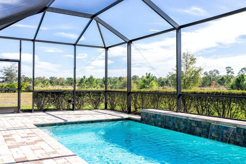 Bright screened-in swimming pool with clear blue water, stone deck, and lush rural landscape beyond under a sunny sky; peaceful retreat.