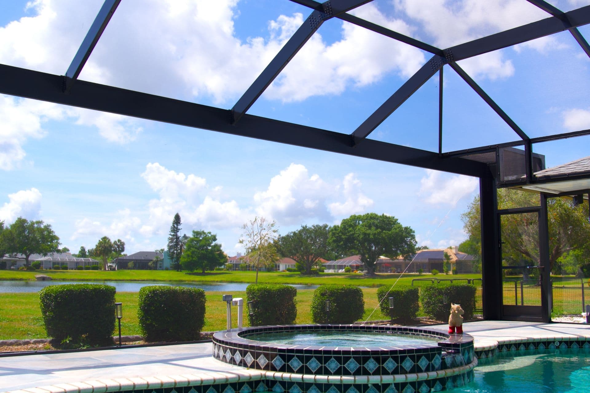 Sunlit screened patio with a tiled spa and swimming pool, manicured hedges, and a peaceful suburban landscape beneath blue, cloudy skies.