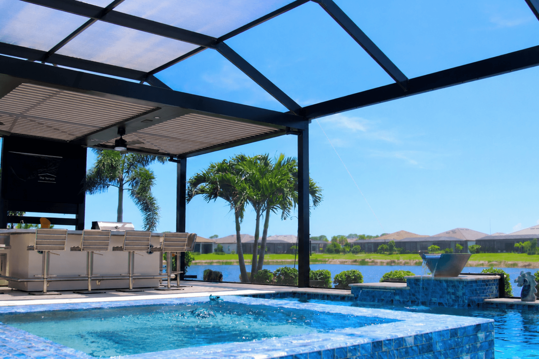 Bright resort pool beneath a screened enclosure, with a pergola, palm trees, lake, and distant houses under a clear blue sky, creating a tranquil mood.