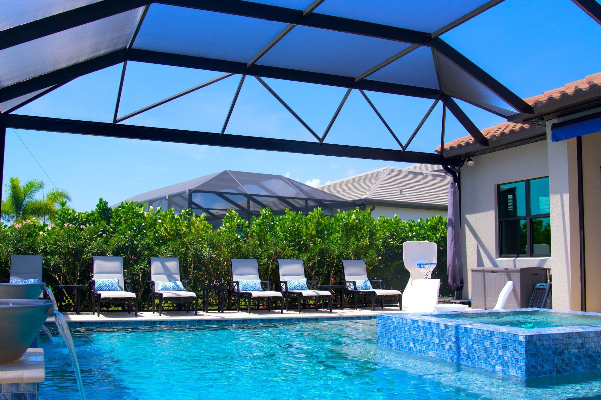 Sunlit screened-in swimming pool beside a modern home, with blue water, white lounge chairs, tropical greenery, and clear sky.