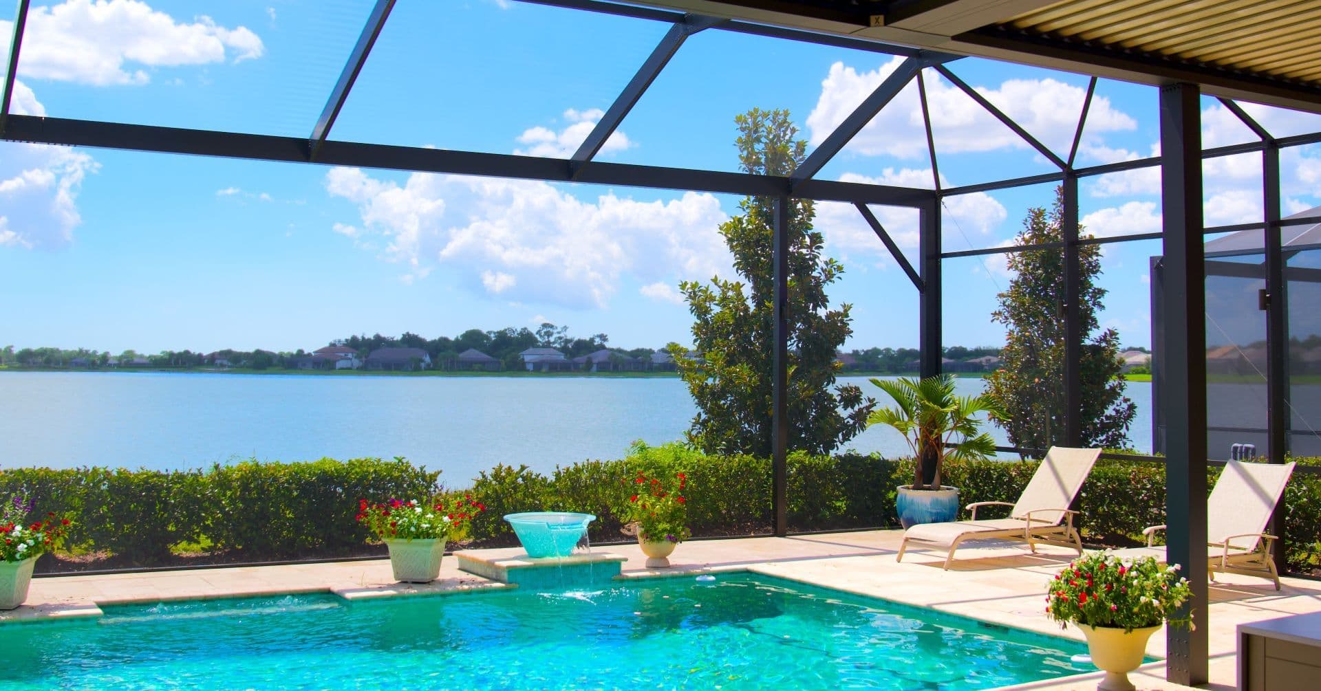 Bright lakeside patio with turquoise swimming pool, lounge chairs, potted flowers, and screen lanai overlooking calm water under a blue sky.