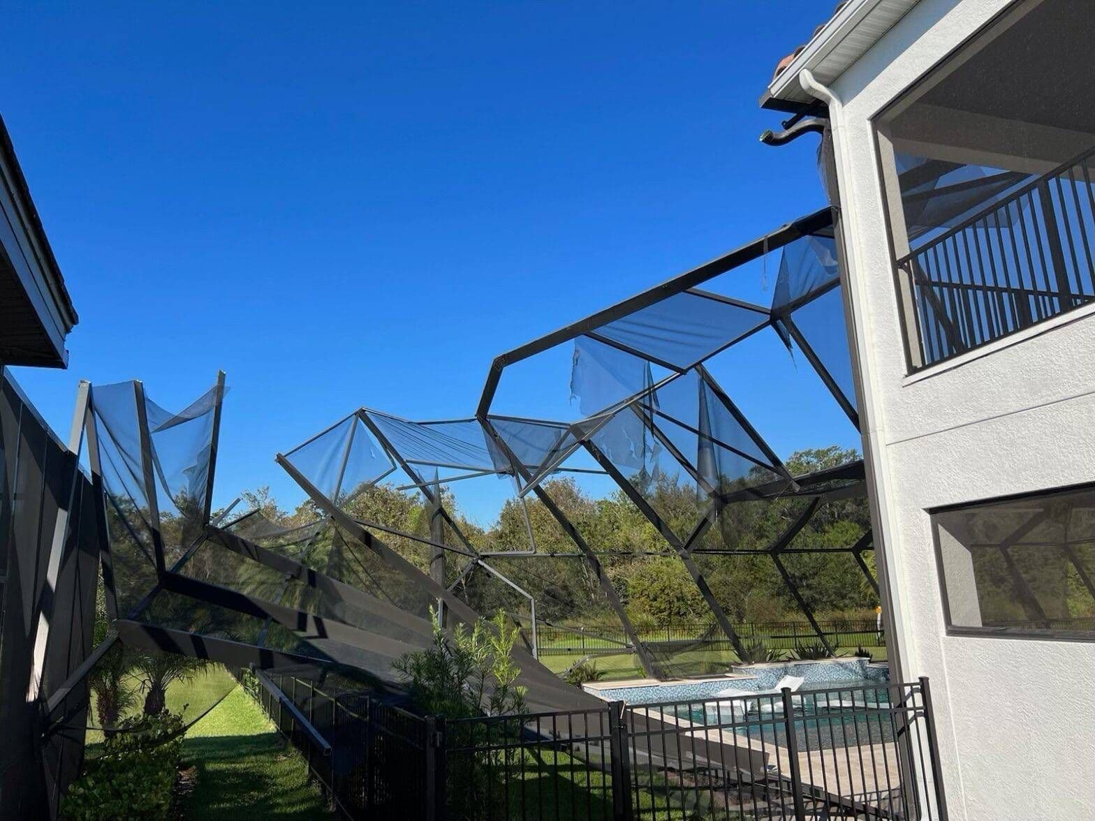 A suburban backyard under a bright blue sky shows a damaged, collapsed pool screen enclosure beside a white house, fence, trees, and pool.