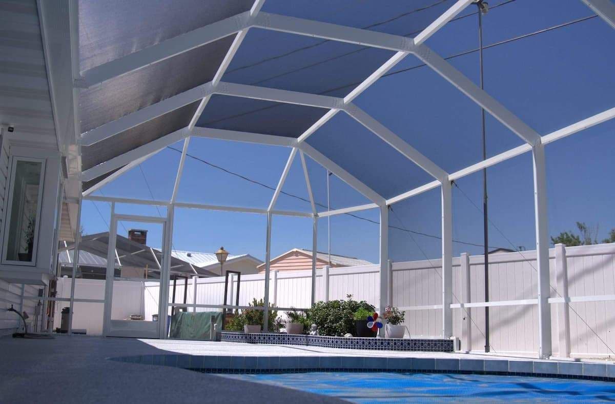 Sunlit screened pool enclosure with white frame, blue swimming pool, potted plants, privacy fence, suburban homes, and clear sky.