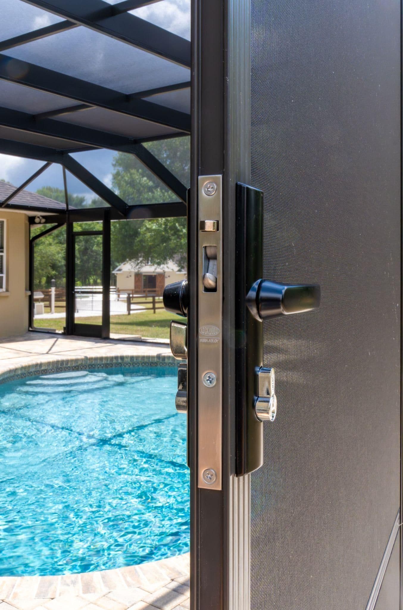 Open black aluminum door with chrome handle leading to indoor pool enclosure with blue water and green outdoor area