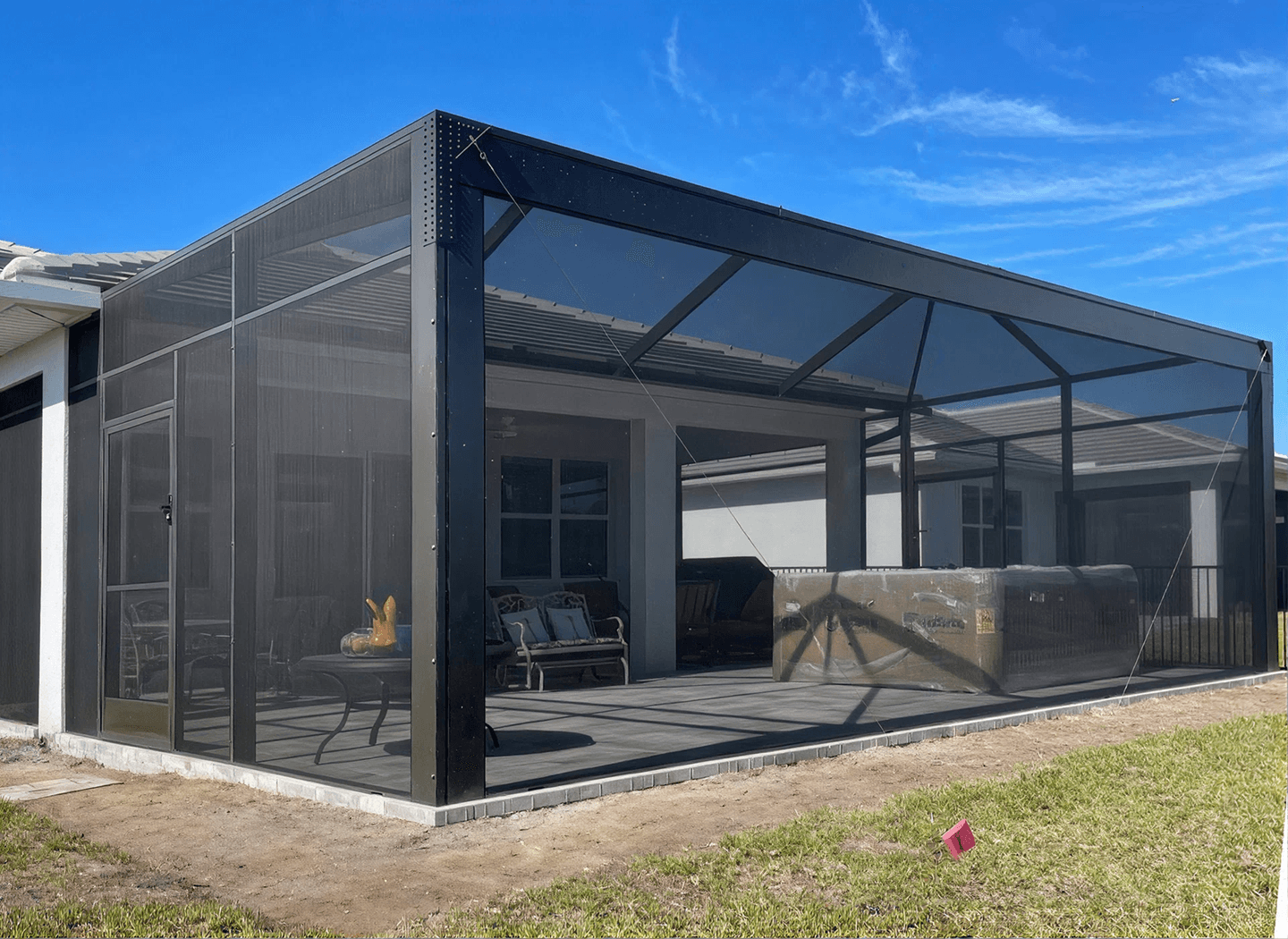 A modern outdoor patio enclosed with a black metal mesh structure, featuring cozy seating and a fireplace, under a bright blue sky.