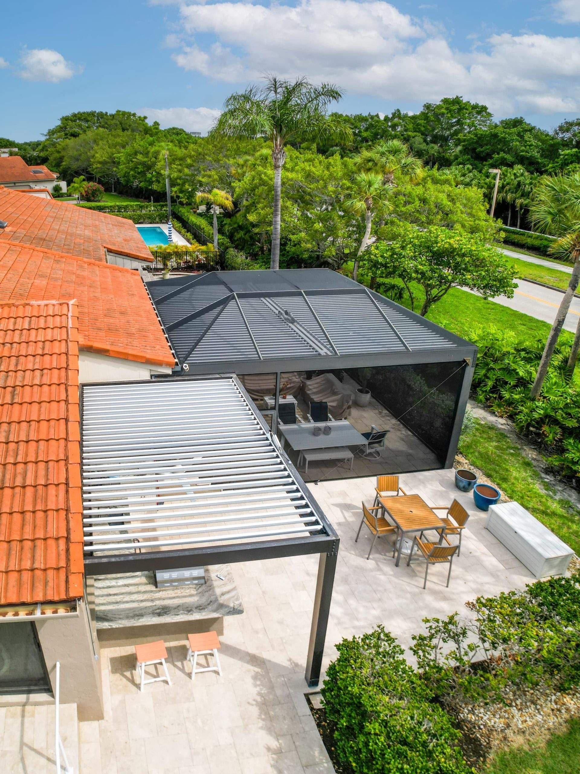 Aerial view of a tropical home with terracotta roofs, covered patio, outdoor dining, lush palms and trees, and a bright blue sky.