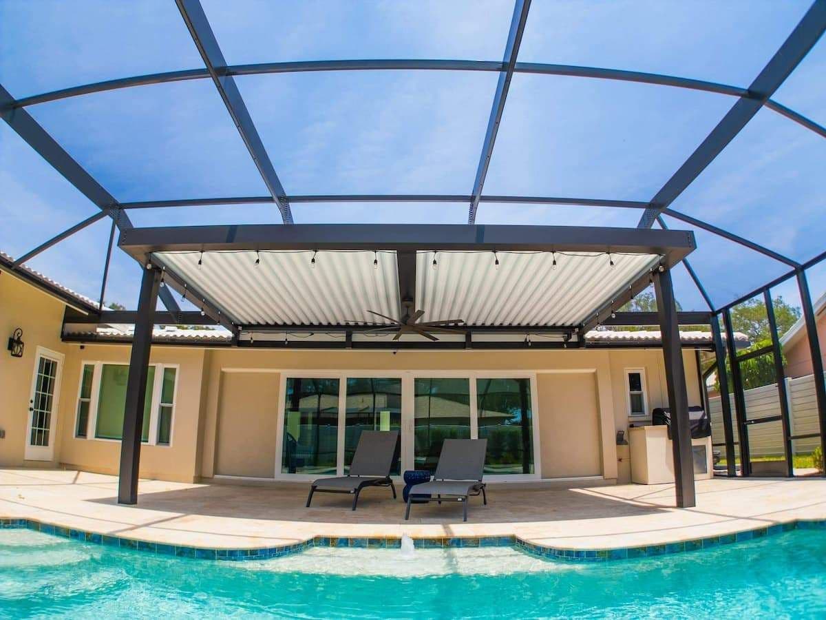 Turquoise backyard pool fronts a tan house with glass doors, shaded by a black pergola; two lounge chairs create a calm, sunny retreat.