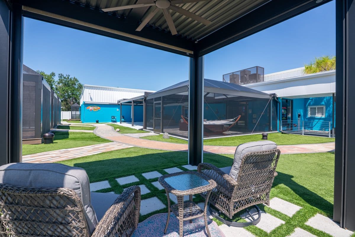 View from covered patio with wicker seating looking out onto a courtyard with blue buildings, green lawn, and brick pathways under clear sky.