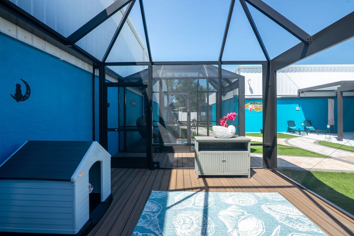 Modern screened patio with blue walls, wooden deck, and sheer roof panels overlooking a lawn with lounge chairs and coastal-themed decor.