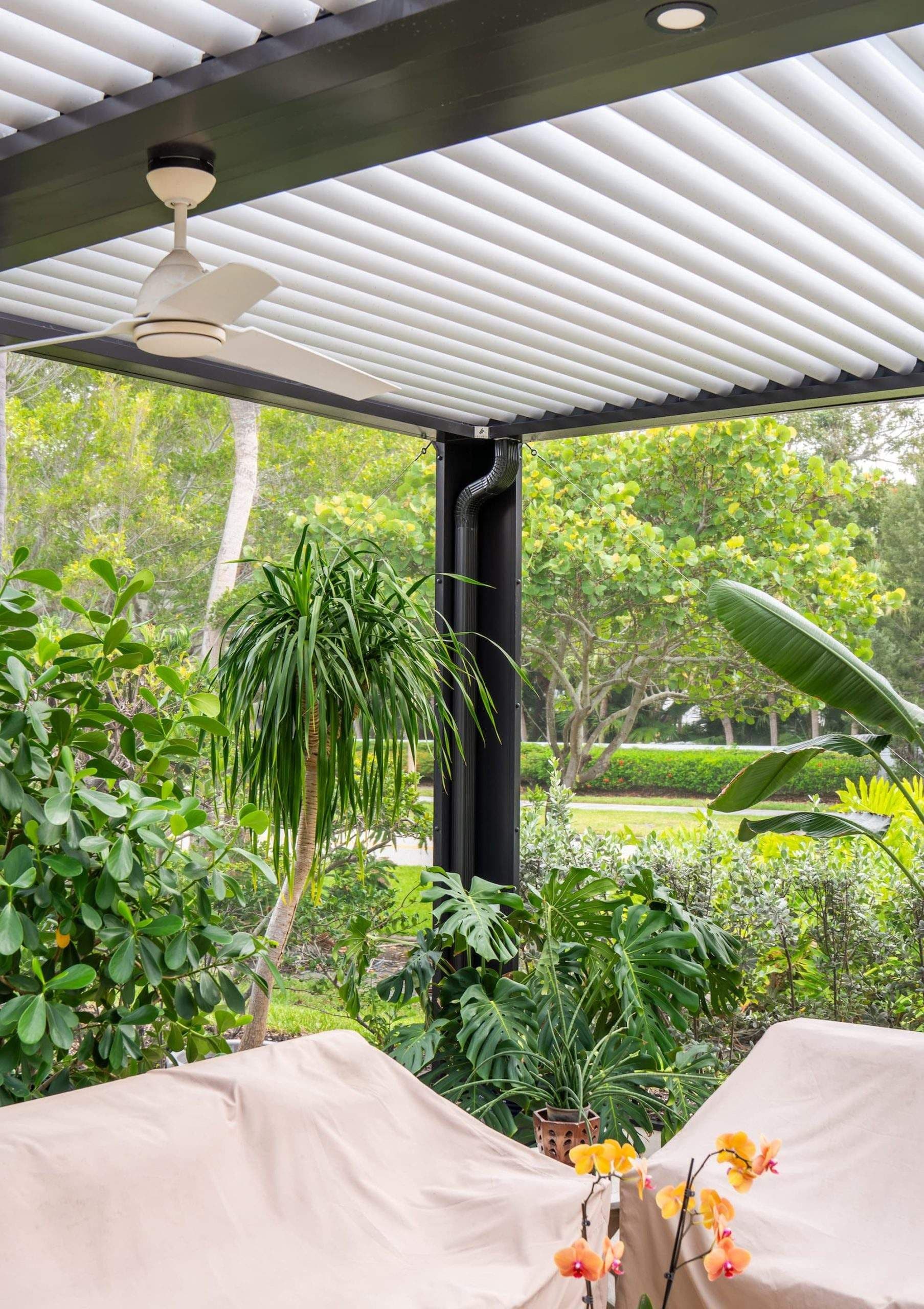 Modern covered patio with white louvered pergola, ceiling fan, potted plants, and view of lush green backyard and mature trees.