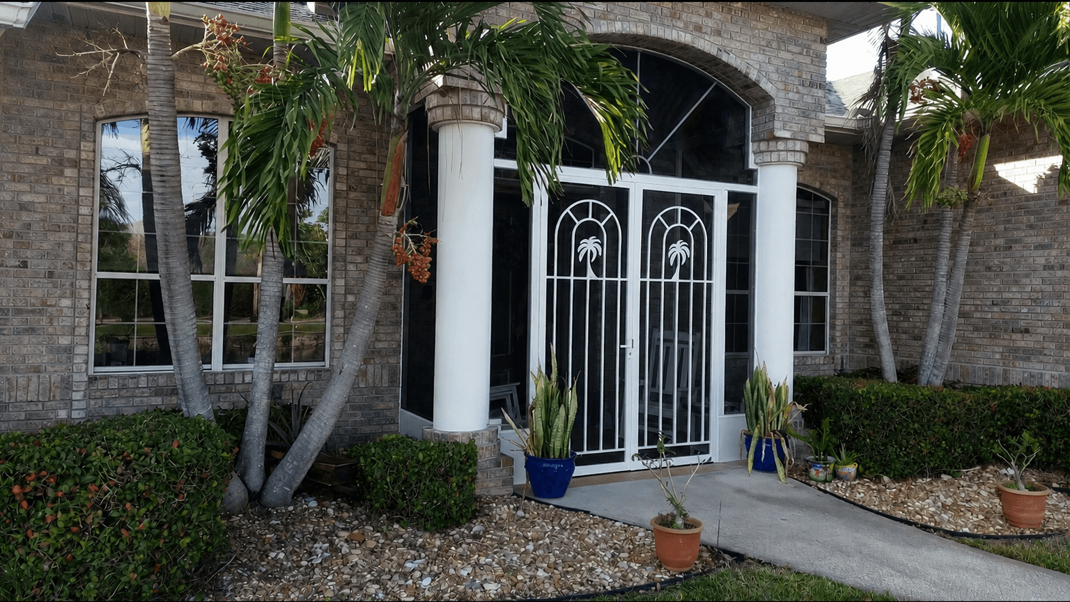 Brick-front tropical home entrance with white columns, ornate double gates, palm trees, potted plants, and a stone-covered garden path.