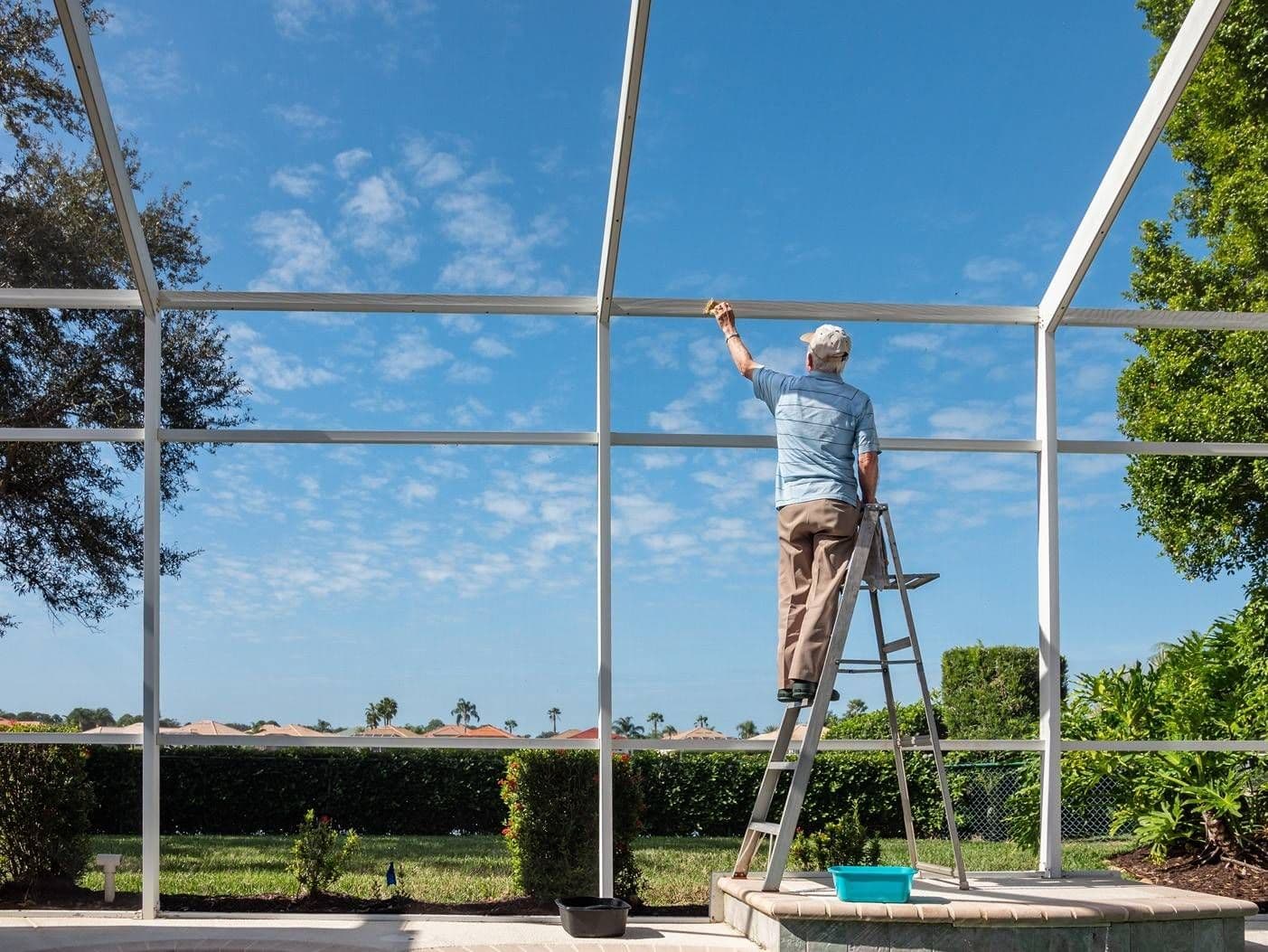 Man on a ladder paints a metal patio enclosure under a bright blue sky, with green hedges, trees, and a sunny backyard creating a calm scene.