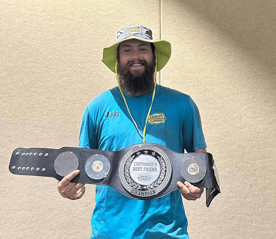 Smiling bearded man in a sun hat and blue shirt holds a large championship belt that reads "Customer's Best Friend", posing proudly against a beige wall.