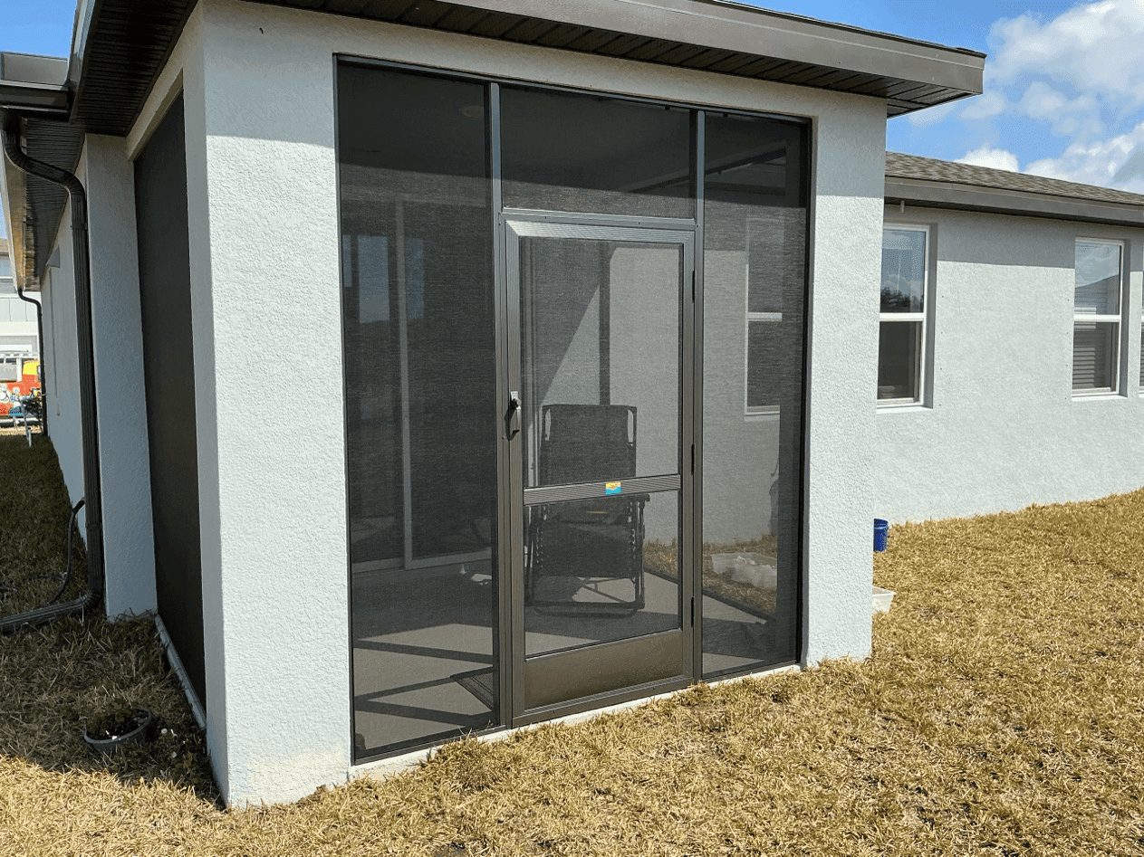 Modern white stucco building with a screened glass entry door, black frames, and dry grass under bright sunlight; clean, quiet exterior.