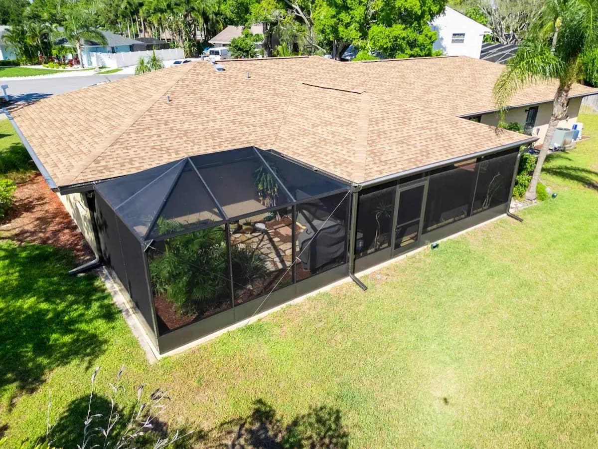Aerial view of a tan-roofed home with a screened-in patio, green lawn, palm trees, and sunny, peaceful suburban surroundings.